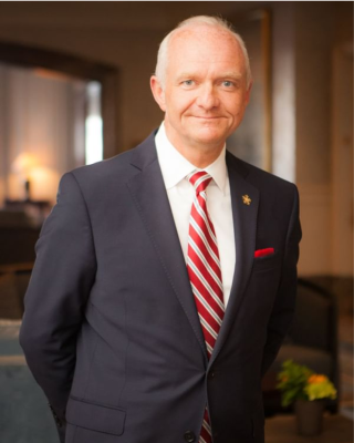 An individual in a suit with a red-striped tie stands indoors, hands behind their back, in a well-lit room that features a plant and some furniture.