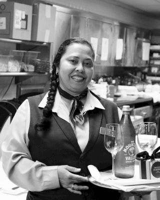 A smiling server in a restaurant holds a tray with a bottle of Moët & Chandon, two glasses, and a corkscrew on a napkin, standing in front of the kitchen.