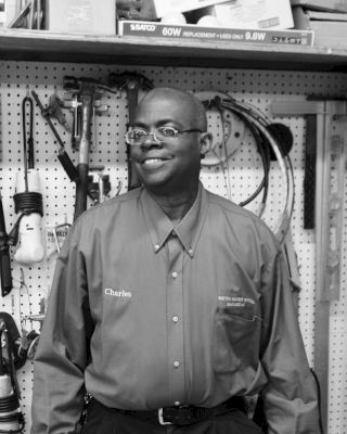 A person wearing a button-up shirt with name embroidery stands in front of a pegboard filled with tools and equipment in a workshop setting.