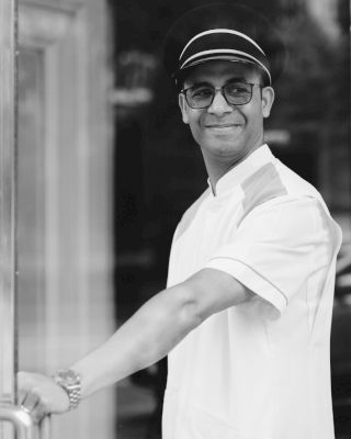 A smiling man wearing a white uniform and cap is holding open a glass door, looking directly at the camera.