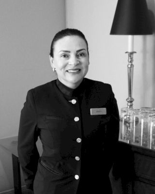 A woman in a uniform is smiling at the camera. She is standing beside a table with several glasses and a lamp on it.