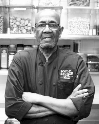 A man in a chef's coat labeled "Boston Harbor" stands with arms crossed, in front of shelves filled with jars of ingredients, facing the camera.