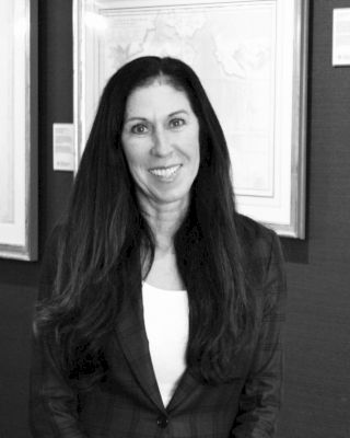 A black-and-white photo of a smiling woman with long hair, standing in front of framed artwork on the wall, dressed in a blazer and white top.