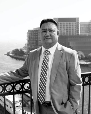 A man in a suit stands on a balcony with a cityscape and waterfront in the background, captured in black and white.