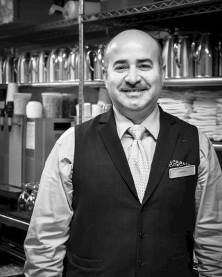 A man is standing and smiling in what appears to be a professional or service environment, dressed in a vest and tie, likely a server or bartender.