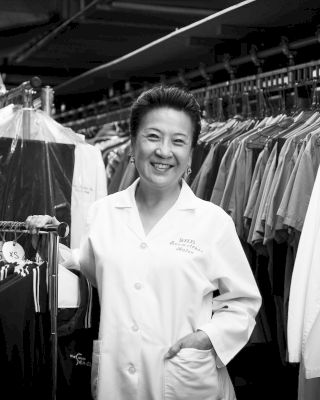 A person in a lab coat standing and smiling, surrounded by rows of hanging garments in what appears to be a dry cleaning facility.