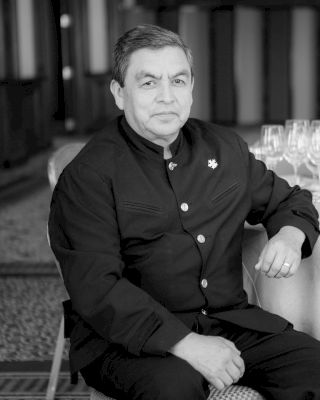 A person dressed in formal attire sits at a table with numerous wine glasses arranged neatly.