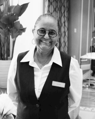 A smiling person wearing glasses and a uniform with a name tag stands indoors, with plants and decorative elements in the background.