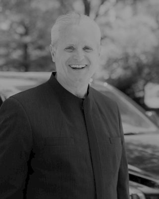 A smiling man in a dark suit stands in front of a vehicle with trees blurred in the background. The image is in black and white.