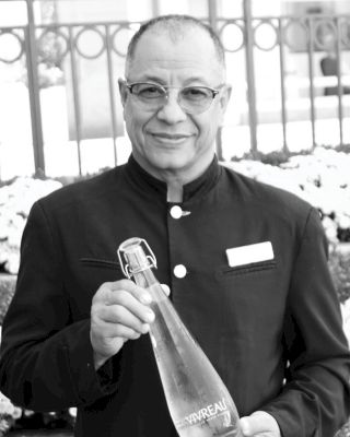 A man in a uniform is holding a bottle of water with a smile, standing outdoors with a fence and plants in the background.