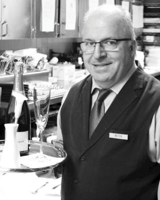 A suited man with glasses, holding a tray with a wine bottle and two glasses, stands in a kitchen-like setting, smiling.