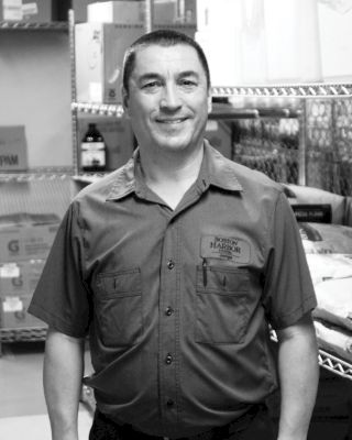 A man in a uniform, smiling, stands in a room filled with boxed and packaged items on shelves.