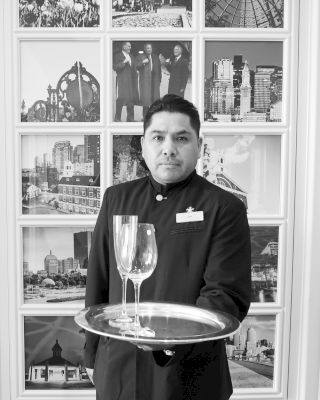 A uniformed person holds a tray with two glasses in front of a wall displaying various framed black-and-white photographs, some showing landmarks.