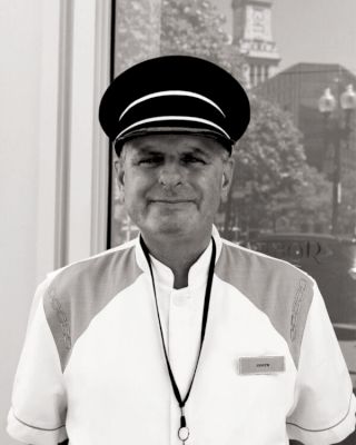 A black and white photo of a man wearing a uniform and a hat, standing and smiling, with a building in the background.