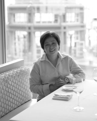 A person smiling and sitting at a table in a restaurant with a building visible through the window in the background.