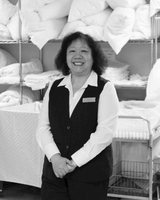 A smiling person in uniform stands in front of linen shelves loaded with folded towels and bedding in a housekeeping or laundry setting.