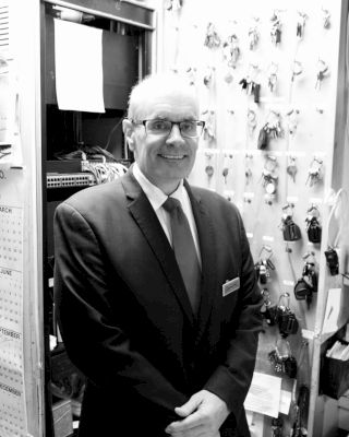 A man in a suit stands smiling in front of a wall with many keys hanging, likely indicating a security or management role.