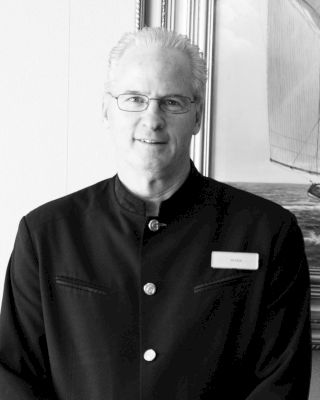 A person in a uniform is standing in front of a framed picture, wearing glasses and a name tag.