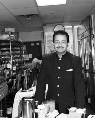 A person in a uniform stands in a kitchen, smiling at the camera. The background features shelves and a catering setup with dishes and cutlery.