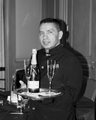 A waiter holding a tray with a bottle of champagne and two champagne glasses. The background features a formal room setting.