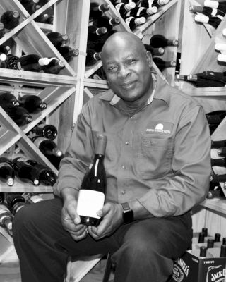 A person is seated in a wine cellar, holding a bottle of wine, with shelves of wine bottles in the background.