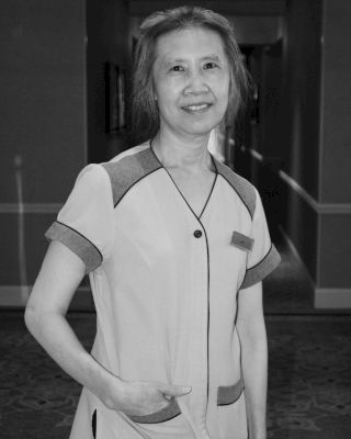 A person is standing in a hallway. They are smiling and wearing attire that might be work-related. The photo is in black and white.