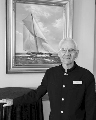 An older man in a black uniform stands in front of a painted sailboat picture, with one hand on a covered table, smiling at the camera.