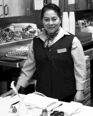 A person is standing in a kitchen or work area with various utensils and items on the counter, wearing a uniform and smiling at the camera.