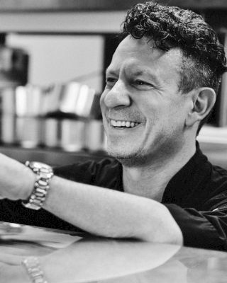 A person is smiling while leaning on a counter in a kitchen environment, with another individual visible in the background.