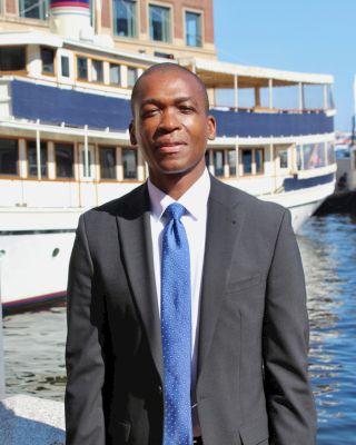 A man in a suit and blue tie stands in front of a boat docked by the water on a sunny day. The background features a large, multi-deck boat.