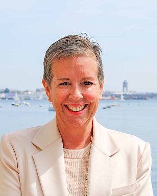 A smiling person in a light-colored blazer stands in front of a scenic waterfront with boats and a distant city skyline, on a clear day.