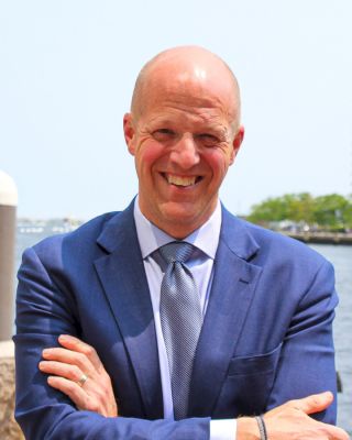 A smiling man in a blue suit with a blue tie stands outdoors near a body of water, arms crossed, with greenery visible in the background.