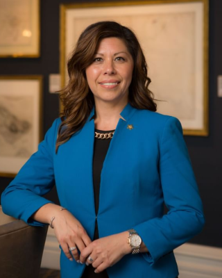 A person is standing in front of framed pictures, wearing a blue blazer, a black top, and a watch, and is smiling at the camera.