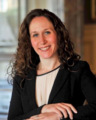 A woman with curly hair is smiling while posing in a professional setting, dressed in a black blazer and white top, hands resting on a table.