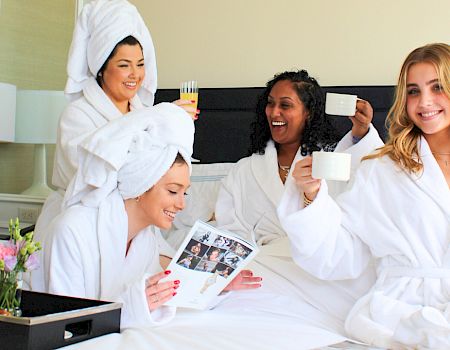 Four women in bathrobes enjoy a relaxing time in a bedroom with drinks, a magazine, flowers, and a tray on the bed, all laughing and smiling.