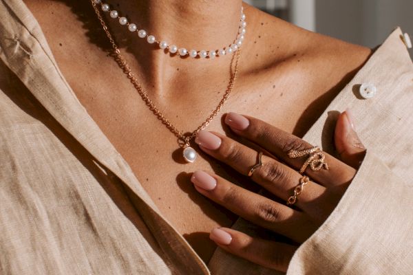 A person wearing a beige shirt, pearl earrings, a pearl choker, and a pendant necklace. Their hand adorned with rings and light pink nail polish.