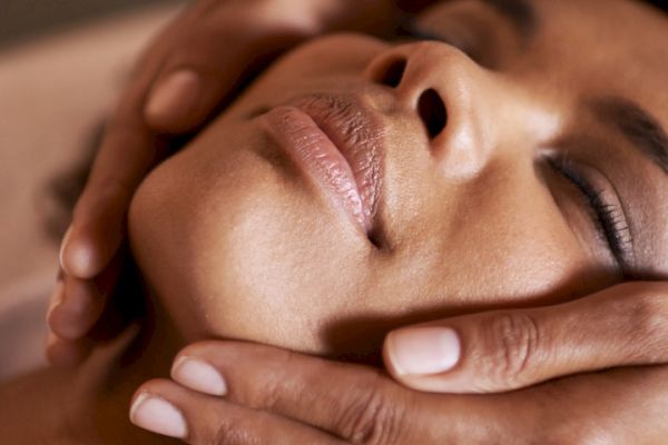 A person receives a gentle facial massage, hands placed on their face and neck, eyes closed in relaxation.