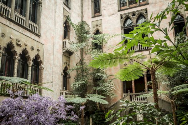 The image shows an old building with arched windows surrounded by lush greenery and purple flowers, creating a serene and historic atmosphere.