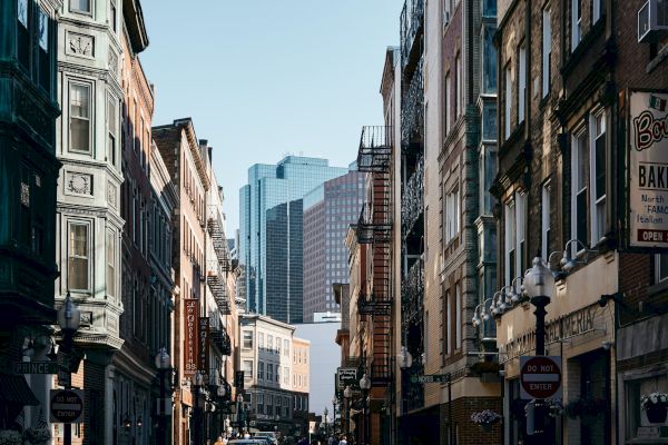 The image depicts an urban street with historic buildings, narrow pathways, and a bakery sign on the right. Modern skyscrapers are visible in the background.