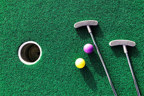 The image shows a mini-golf hole on green turf, with two putters and two golf balls, one yellow and one purple, placed near the hole.