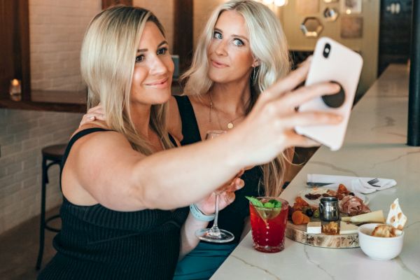 Two women are taking a selfie at a bar. One holds a drink, and there are various snacks and drinks on the table.