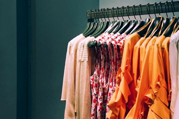 A row of colorful clothes, including white, patterned, and orange outfits, neatly hang on a metal clothing rack in a retail store.