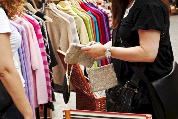 Two people are at an outdoor clothing market; one holds a beige clutch, while various colorful garments hang on a rack in the background.