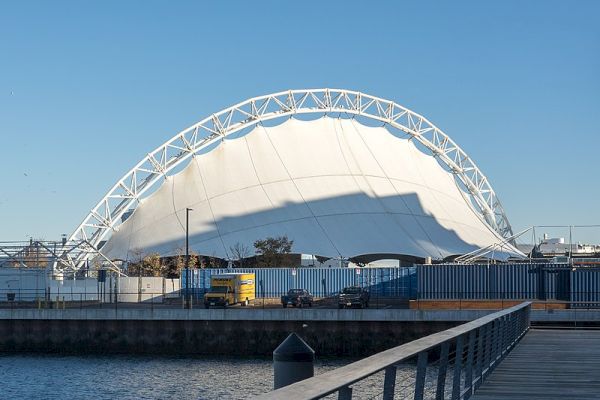 This image features a large, white, dome-shaped structure with a metal framework, situated near a waterfront, alongside buildings and vehicles.