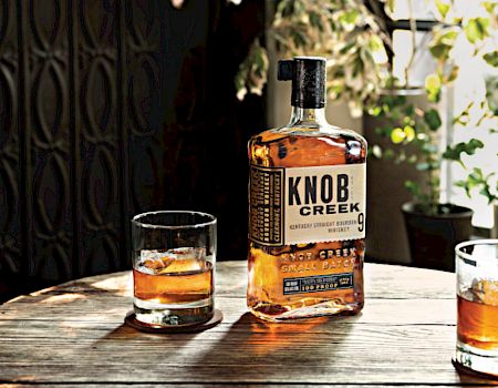 The image shows a bottle of Knob Creek whiskey and two glasses with whiskey on a wooden table, surrounded by a cozy, plant-filled setting.