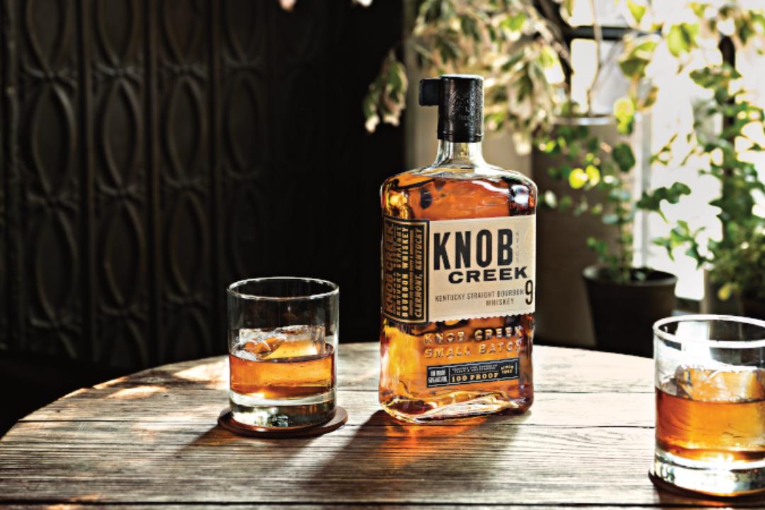 The image shows a bottle of Knob Creek whiskey and two glasses with whiskey on a wooden table, surrounded by a cozy, plant-filled setting.