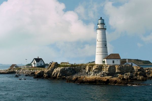 A lighthouse on a rocky island with adjacent small buildings and a cloudy sky in the background sits surrounded by water.