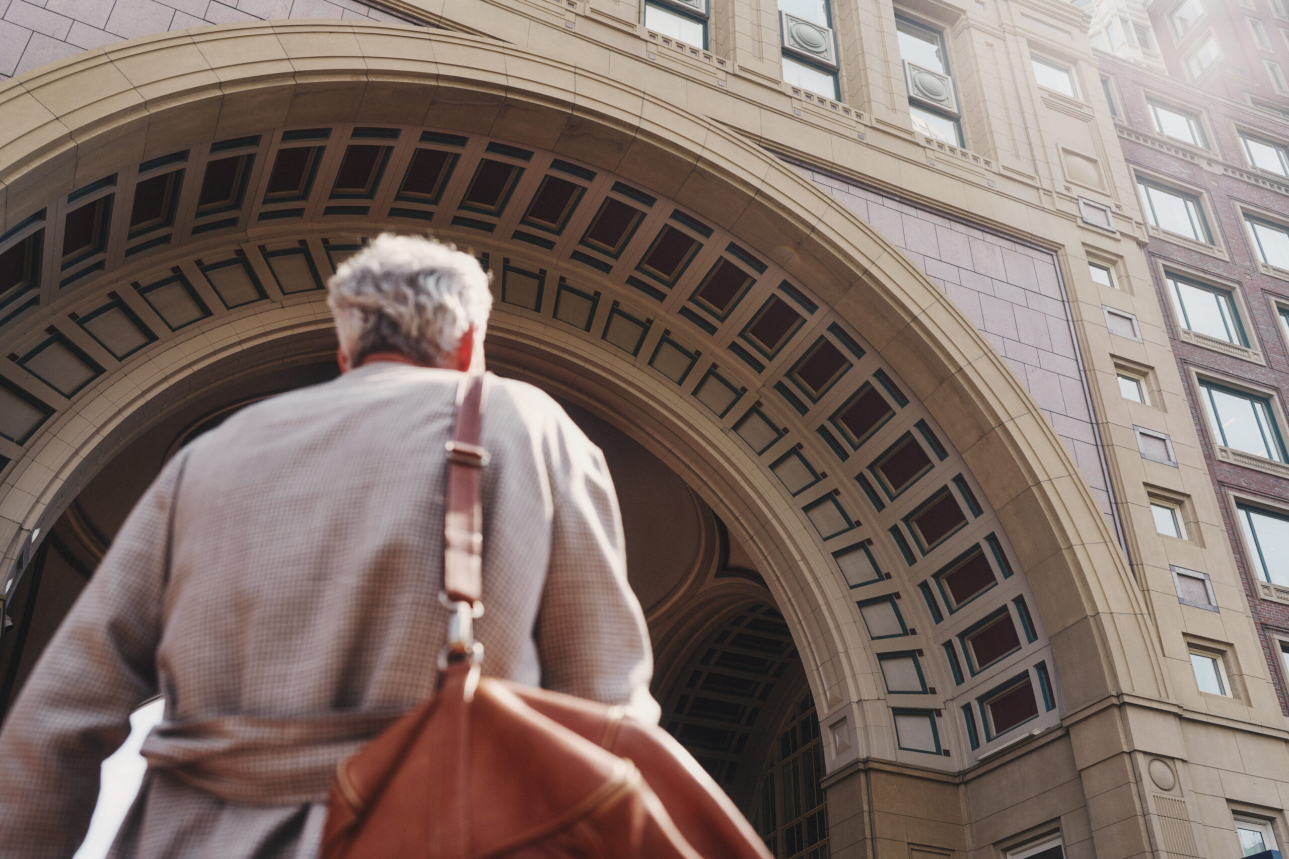 A person with gray hair and a brown bag is walking towards an arched architectural structure made of stone, visible from a low angle.