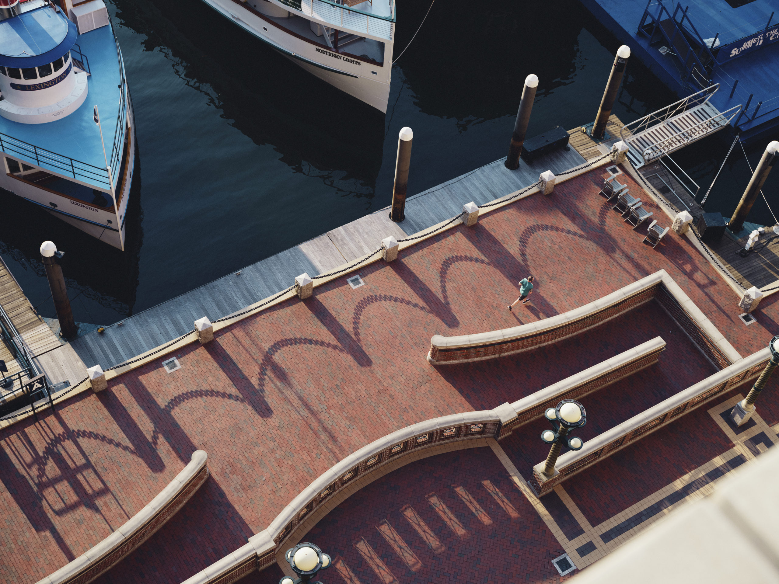 An aerial view of a waterfront promenade with boats docked at the pier, light poles casting shadows, a lone walker, and decorative brickwork.