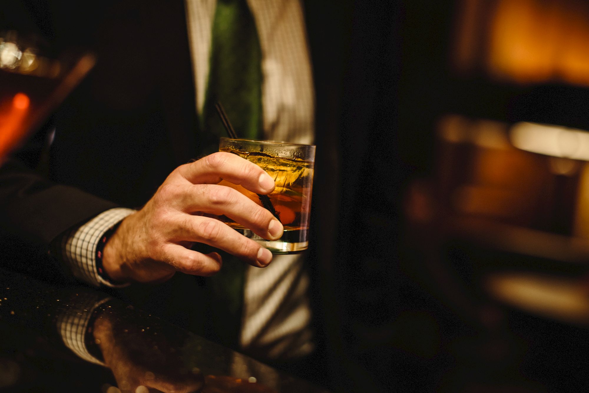 A person in a suit holds a glass of whiskey with ice at a dimly lit bar.